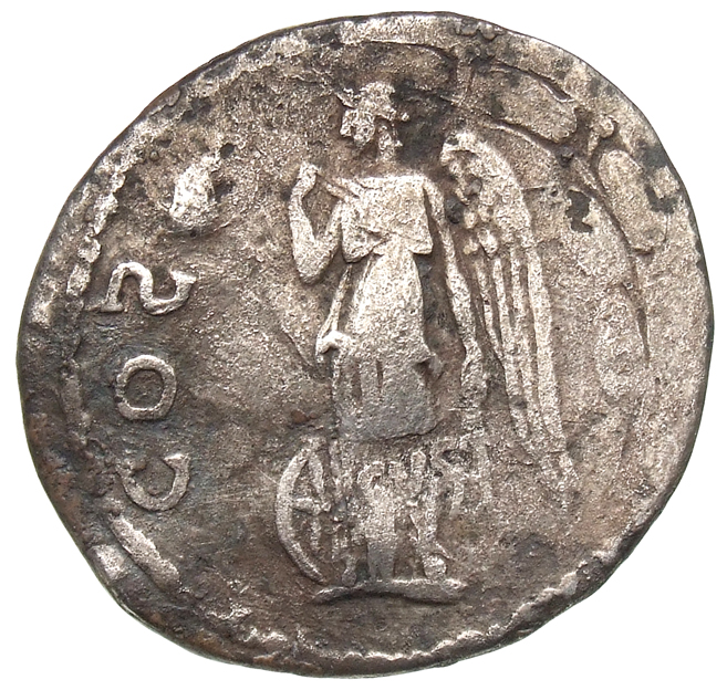 Hadrian reverse — Geflügelte Nemesis steht in der Vorderansicht, den Kopf nach l. Sie greift mit ihrer r. Hand in das Gewand und hält in der l. Hand die Zügel. Hinter ihr l. ein Rad. Im l. F. Reste des Untertyps mit Fischschwanz und Füllhorn des Capricorn.